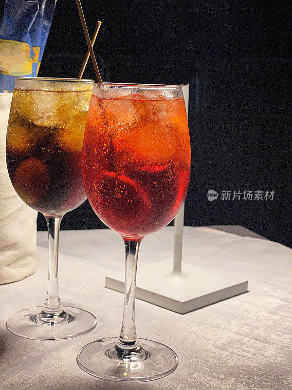 餐厅的桌子上放着几杯Hugo和Cynar Spritz鸡尾酒，用木棍装饰着绿橄榄，夜晚的户外用餐环境，重点放在前景